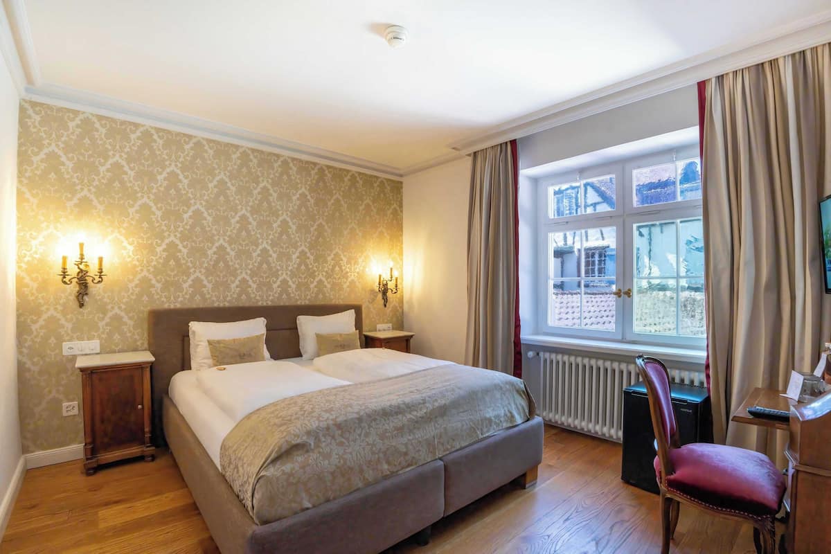 A well-lit bedroom with a large bed, two bedside tables, a chair, and a view of a building through a window.