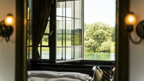 Une chambre avec un lit, une fenêtre avec des rideaux et une vue sur un paysage bordé d’arbres.