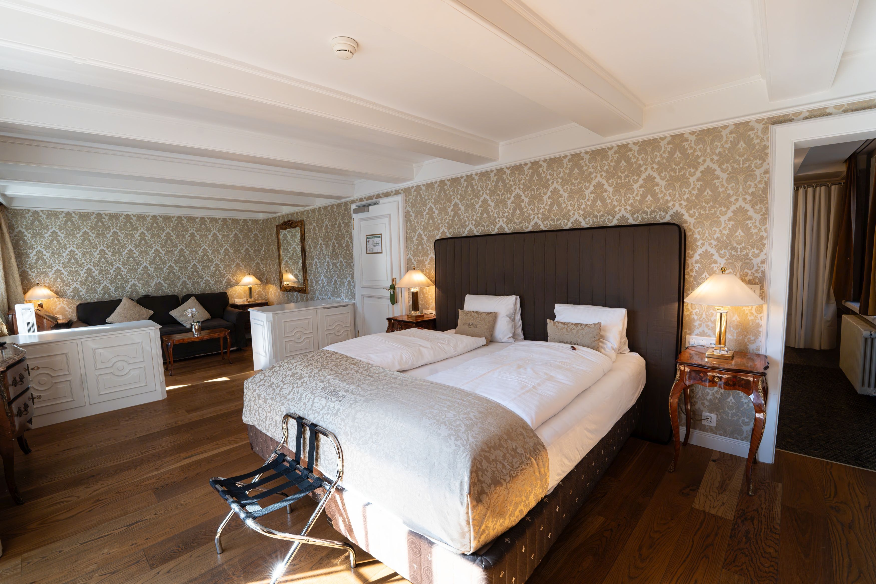 Un dormitorio amplio con una cama grande, una zona de estar con sofá y sillones, piso de madera y un espejo decorativo.