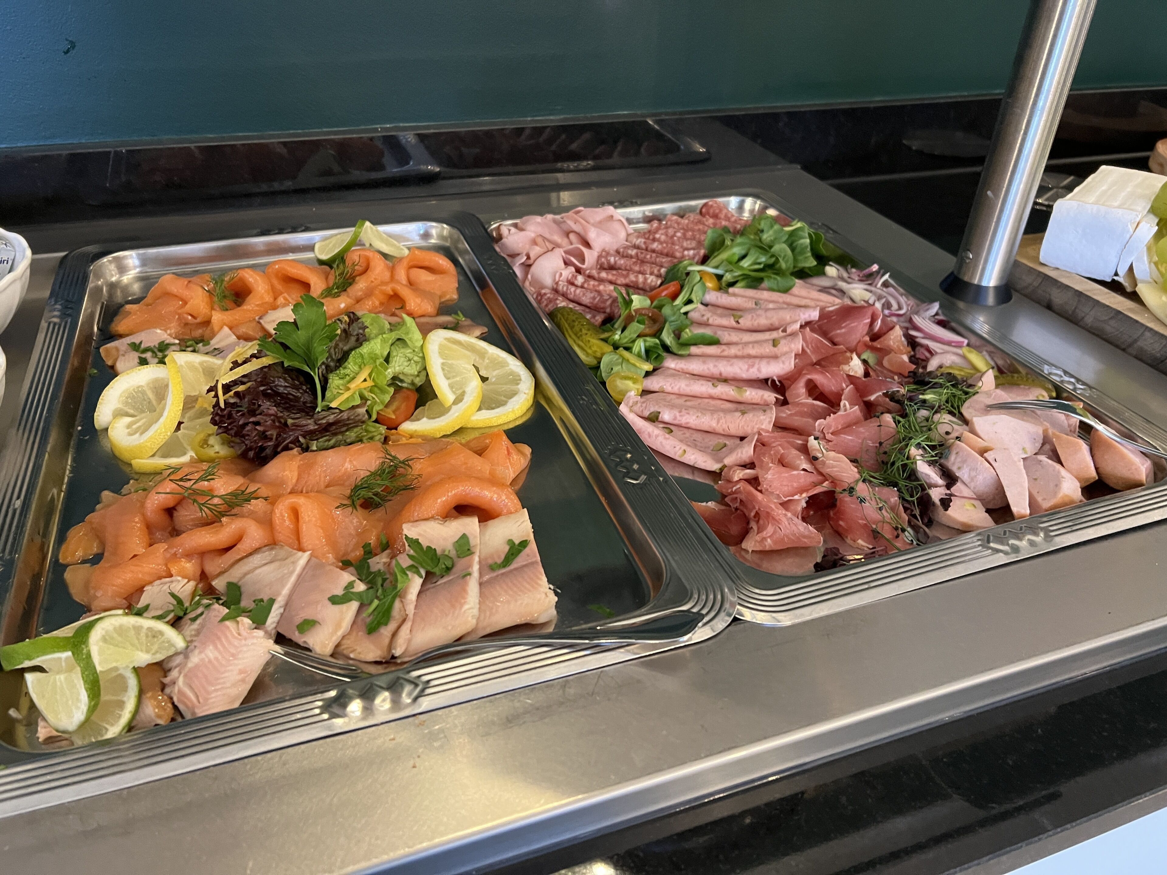 A buffet with various types of deli meats and smoked salmon, garnished with herbs and lemon slices.