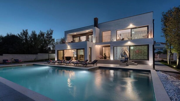 Une maison moderne à deux étages, dotée d’une piscine, de chaises longues et d’une grande fenêtre au motif géométrique.