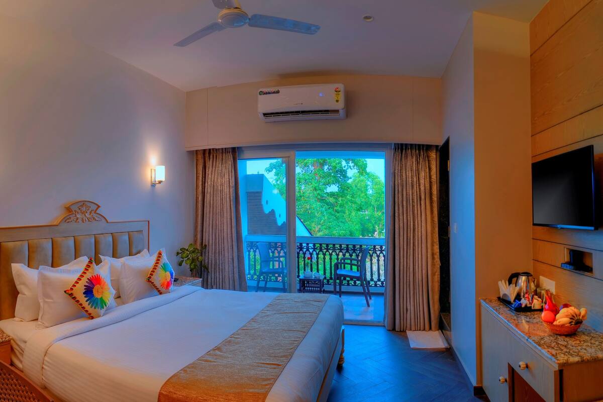 Habitación de hotel con cama, escritorio, televisión y balcón con vistas a árboles.