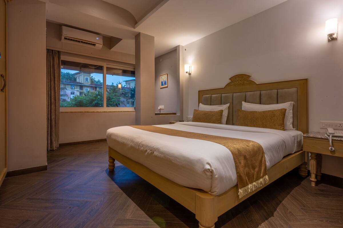 Una habitación de hotel con una cama grande, una mesita de noche con lámpara, un equipo de aire acondicionado, una ventana con cortinas y vistas a edificios.