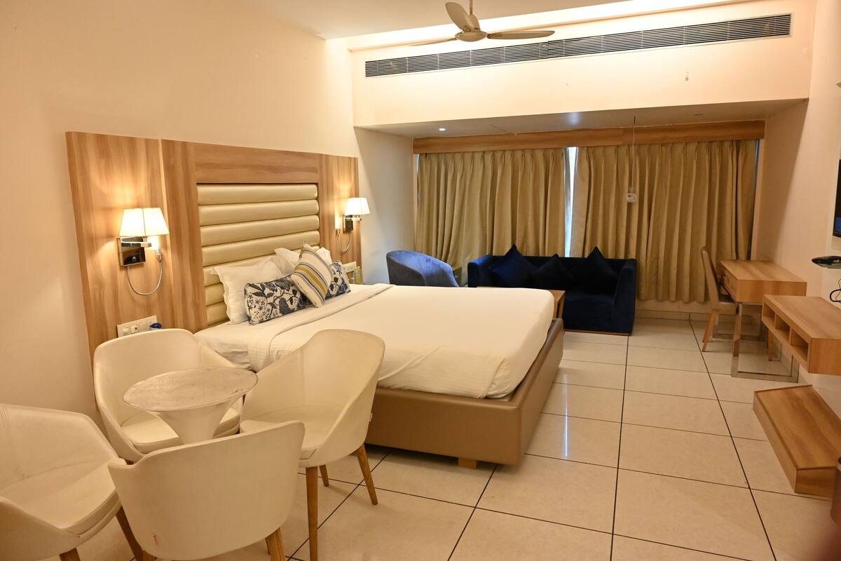 Quarto de hotel com uma cama grande, uma mesa, duas cadeiras, um ventilador de teto e uma televisão.