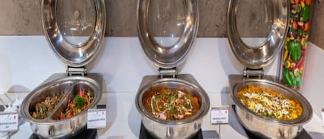 Three buffet bowls with lids open, displaying different dishes, and a variety of utensils on the table.