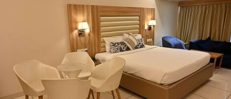 A hotel room with a large bed, two bedside tables, a desk with chairs, and a blue armchair.