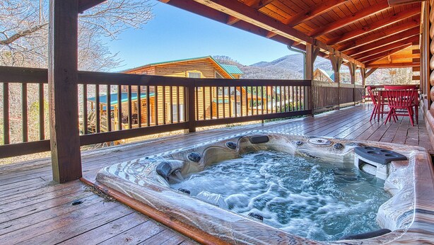Una terraza de madera con un jacuzzi, barandas de madera y vistas a cabañas al fondo.