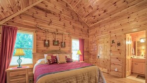 Un dormitorio de cabaña de madera con una cama grande, dos mesitas de noche y un ventanal con cortinas.