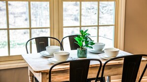 Une table à manger avec quatre chaises, une plante en pot et quatre bols blancs posés sur des assiettes, le tout placé devant une fenêtre donnant sur des arbres.
