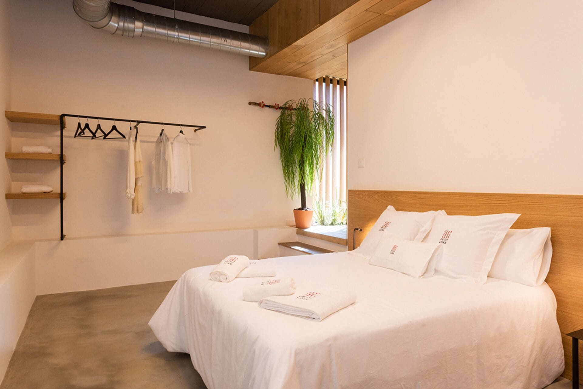 Un dormitorio con cabecera de madera, cama con ropa blanca, planta en maceta junto a la ventana y toallero con toallas.