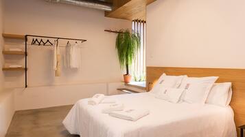 Un dormitorio con cabecera de madera, cama con ropa blanca, planta en maceta junto a la ventana y toallero con toallas.