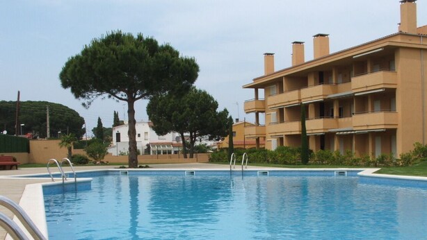 A swimming pool with a clear blue water surface, surrounded by a building with balconies and a tree in the background. - Modern Apartment - Gardens/Pool/WIFI/AIRCON/PARKING - D302, Apartment Riells Blau D302 in L'Escala -... (L'Escala)