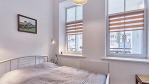 Ein Schlafzimmer mit Bett, Fenster mit Jalousien, einem Bild an der Wand und einem Stuhl.
