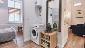 Ein kompakter Wohnraum mit Waschmaschine, Bücherregal, großem Spiegel und einer Sitzecke mit dunkelgrauem Sofa.