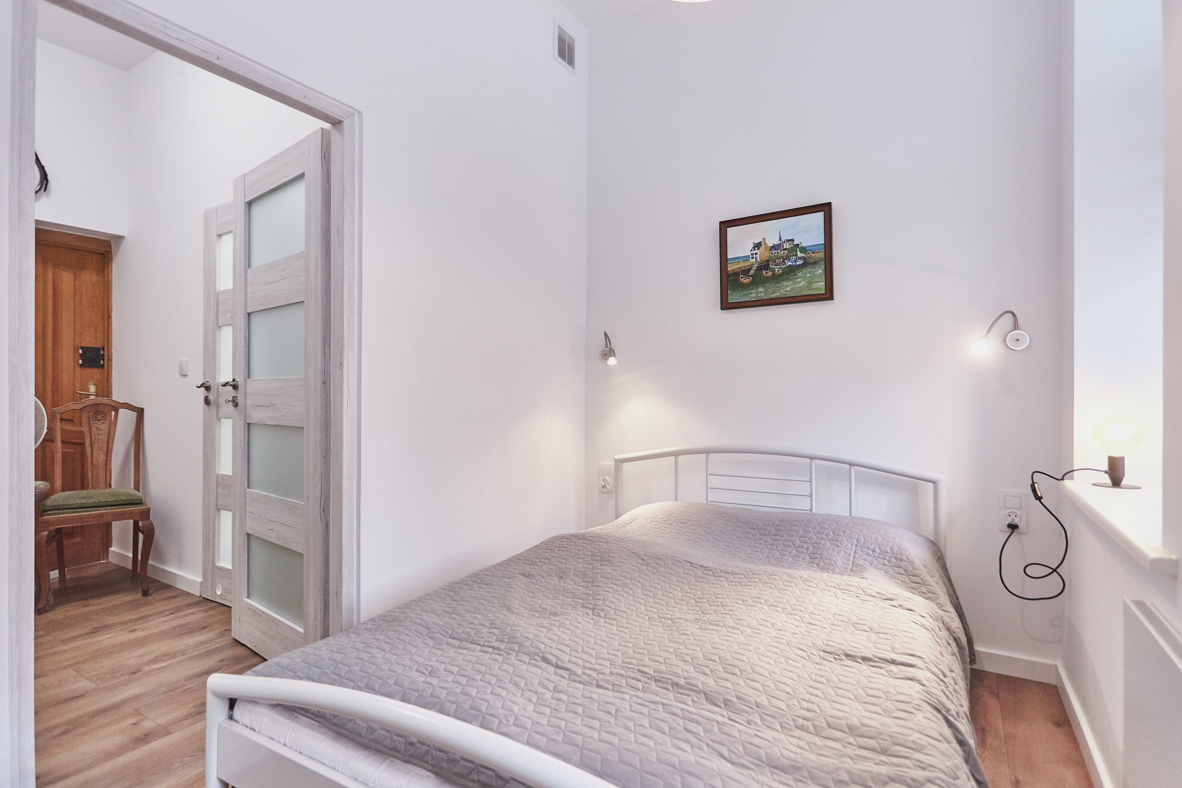 Une chambre à coucher avec un lit, une chaise en bois, une porte blanche dotée de panneaux en verre dépoli, un tableau accroché au mur et une table de chevet avec une lampe.
