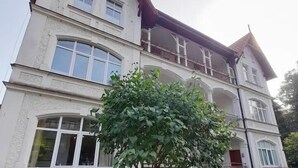 Ein mehrstöckiges Gebäude mit einer symmetrischen Fassade, großen Fenstern und einem Baum im Vorgarten.