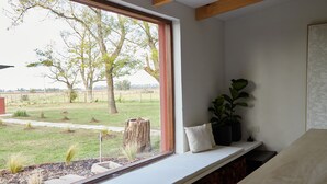 Ein Zimmer mit großem Fenster mit Blick auf eine Wiese mit Bäumen und einem Steinsockel. Eine weiße Bank mit Kissen und zwei Blumentöpfe stehen auf einem Sims.