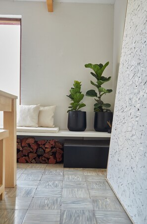 Una habitación con un banco blanco, dos plantas en macetas, una pila de leña y un pared con textura.