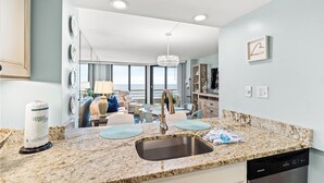 A modern kitchen with granite countertops, a stainless steel sink, and a dishwasher. There is a dining area with a table and chairs, and a large window offering a view of the ocean. - Recently Renovated Oceanfront Condo, Private Resort +Free Attraction Tickets! (Myrtle Beach)