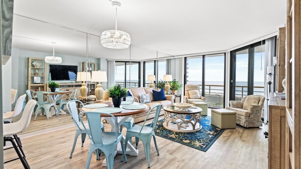A spacious living room with a large window, a dining area with light blue chairs, and a cozy seating area with a sofa and armchairs. - Recently Renovated Oceanfront Condo, Private Resort +Free Attraction Tickets! (Myrtle Beach)