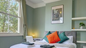 A neatly arranged bedroom with a large bed, bedside tables, a nightstand, a framed picture on the wall, and a window with curtains. - Beautiful, light & spacious Regency 2 bed apt (Royal Leamington Spa)