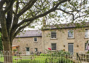 A stone building with multiple windows and a door, surrounded by a metal fence and greenery. - Dale End Farm Cottage (Bakewell)