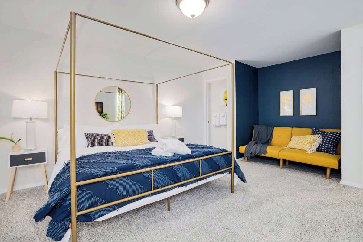 Una cama con dosel con un edredón blanco y azul, una mesita de noche con una lámpara, un sofá amarillo y una pared de acento azul oscuro.