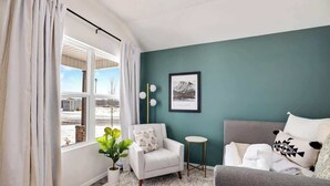Una sala de estar con un sofá, un sillón y una mesita auxiliar. Hay una ventana con cortinas, una planta y un cuadro en la pared.