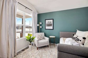 A living room with a sofa, armchair, and a small side table. There is a window with curtains, a plant, and a framed picture on the wall.