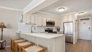 Una cocina con gabinetes blancos, una isla central con taburetes, microondas, refrigerador y cafetera.