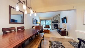 A spacious dining area with a long wooden table, benches, and a large mirror above. A cozy seating area with a fireplace and a fan is visible in the background. - Bright Unit, Quiet Location, Near Mountain Bike Trails, Balcony. On-site Pool (Mammoth Lakes)