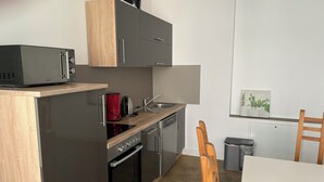 Eine kompakte Küche mit Mikrowelle, Wasserkocher und Backofen. Es gibt ein Spülbecken, einen Stuhl und einen Mülleimer.