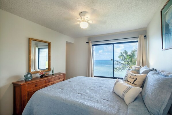 A bedroom with a large bed, a wooden dresser, a mirror, and a window with a view of the ocean.