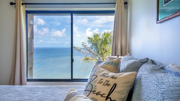 A bedroom with a large window overlooking the ocean, a bed with multiple pillows, and a plant on the windowsill. - Newly updated w/ Tons of Personality & Awesome Oceanfront Location (Princeville)