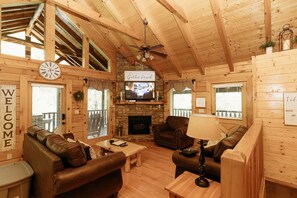 Cozy cabin-style living room with a high ceiling and exposed beams, featuring a mounted TV above a stone fireplace and comfortable leather sofas.