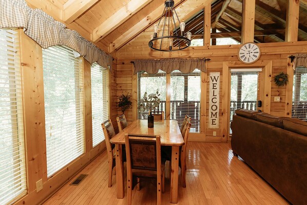 Cozy interior with a high ceiling, wooden beams, and a dining table for four, accompanied by a sofa and large windows with blinds.