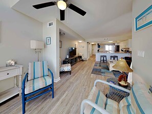 Sala de estar con sofá, sillón y televisor. Se observa una cocina en el fondo con mesa y sillas.