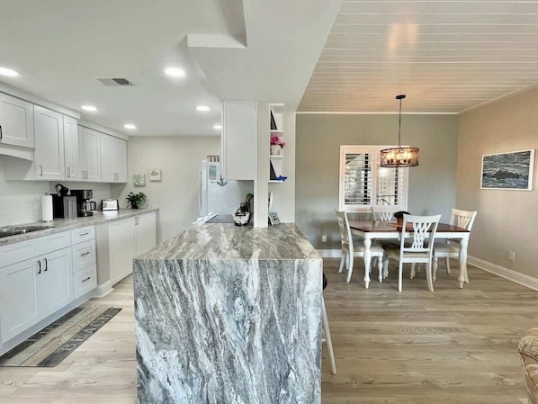 Cozinha moderna com armários brancos, ilha de mármore e sala de jantar com cadeiras brancas e um lustre.