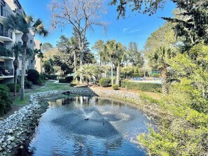 Um lago tranquilo com uma fonte, rodeado por vegetação exuberante e um jardim bem cuidado.