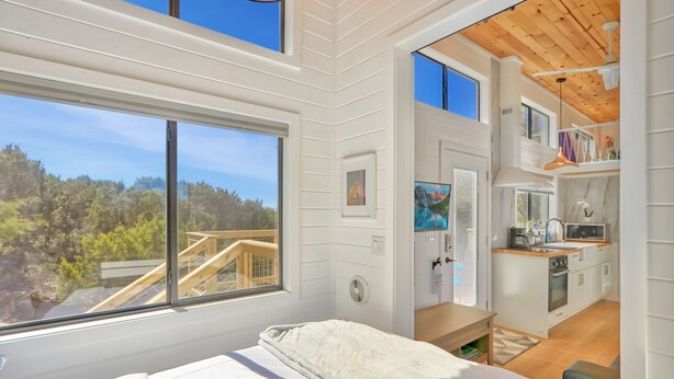 A modern bedroom with a large window, a view of a wooden deck, and a small kitchenette. - Starlight Casita | Modern Tiny House (Fischer)