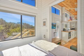Une chambre moderne dotée d’une grande fenêtre, offrant une vue sur une terrasse en bois, et comprenant une petite kitchenette.