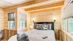 Quarto de cabana de madeira com uma cama, duas janelas com persianas, uma mesinha de cabeceira e um abajur fixado na parede.
