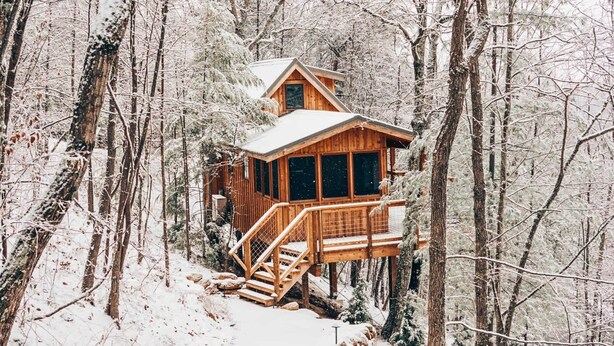 Uma cabana de madeira com varanda e escadas, rodeada por árvores cobertas de neve.