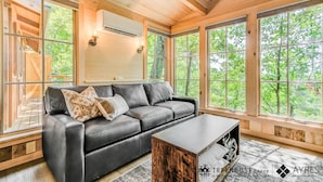 A modern log cabin interior with a leather sofa, a coffee table, and large windows offering a view of the forest.