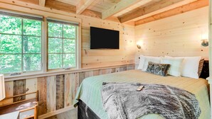 Een slaapkamer in een houten hutje, met een bed, een aan de muur bevestigde televisie en een raam met uitzicht op bomen.