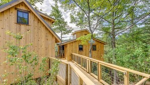 A wooden cabin with a deck and a small attached structure, surrounded by trees. - Accessible 4Bed Treehouse + Kitchen + TV | Spruce (Sevierville)
