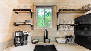 Een compacte keuken met een raam, planken met servies, een koffiezetapparaat en keukengerei.