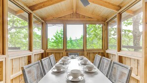 Een eetruimte met een lange tafel voor zes personen, houten wanden en grote ramen met uitzicht op bomen.