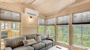 Een gezellig interieur van een blokhut met een grijze bank, een kleine tafel en een raam met uitzicht op het bos.