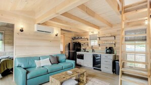A cozy cabin interior with a teal sofa, wooden ceiling beams, a kitchenette, and a large window. - 3Bed Treehouse + HotTub + DogFriendly | Moosewood (Sevierville)
